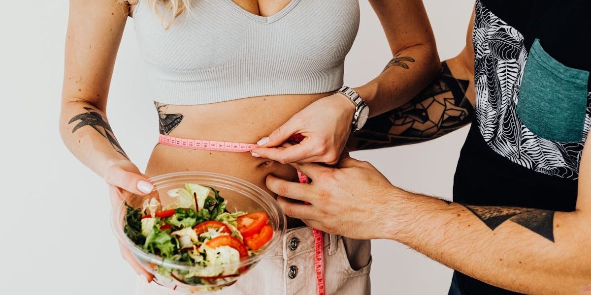 A couple measuring waistline with a tape and holding a fresh salad bowl, focusing on fitness and weight loss.