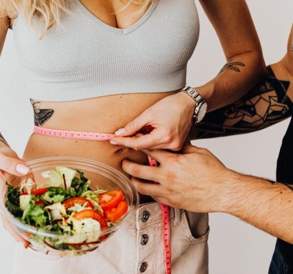 A couple measuring waistline with a tape and holding a fresh salad bowl, focusing on fitness and weight loss.